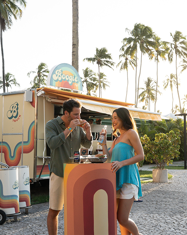 Casal se divertindo no food truck Boogie do Japaratinga Lounge Resort, comendo e bebendo ao ar livre, rodeado por palmeiras.