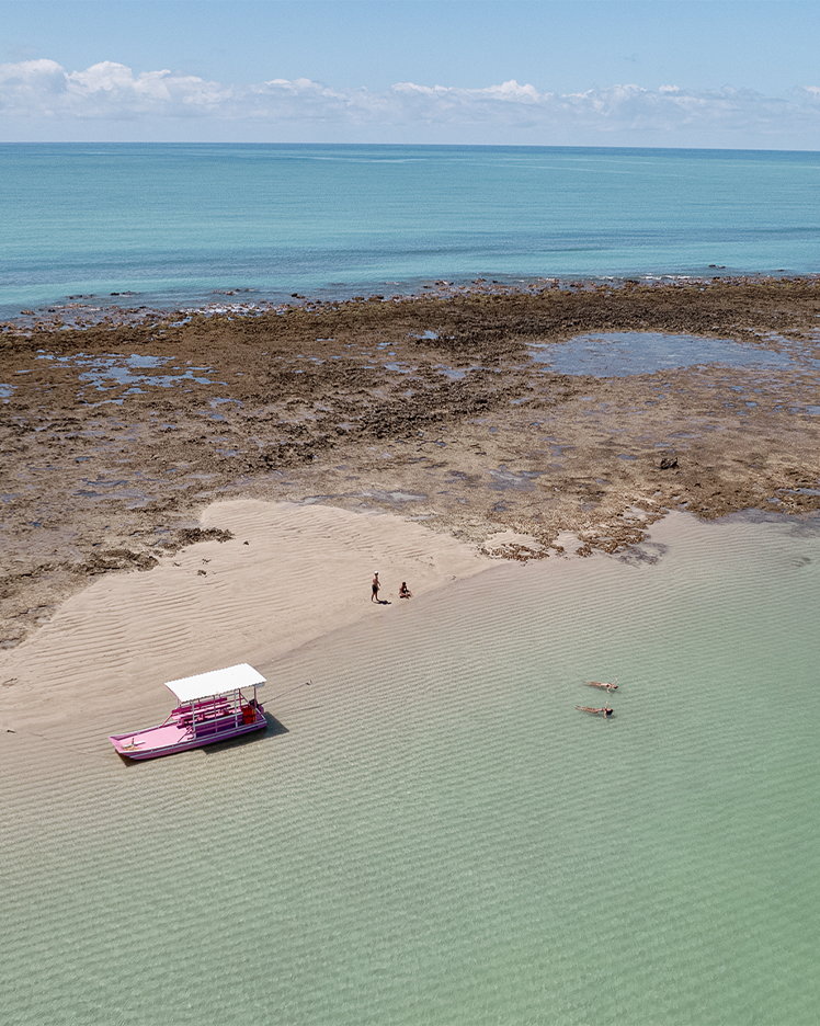 Visto de cima um barco com casal caminhando no raso da Croa de São Bento, com água em tom turquesa e céu sem nuvens. Ao fundo se vê a vegetação de Alagoas.