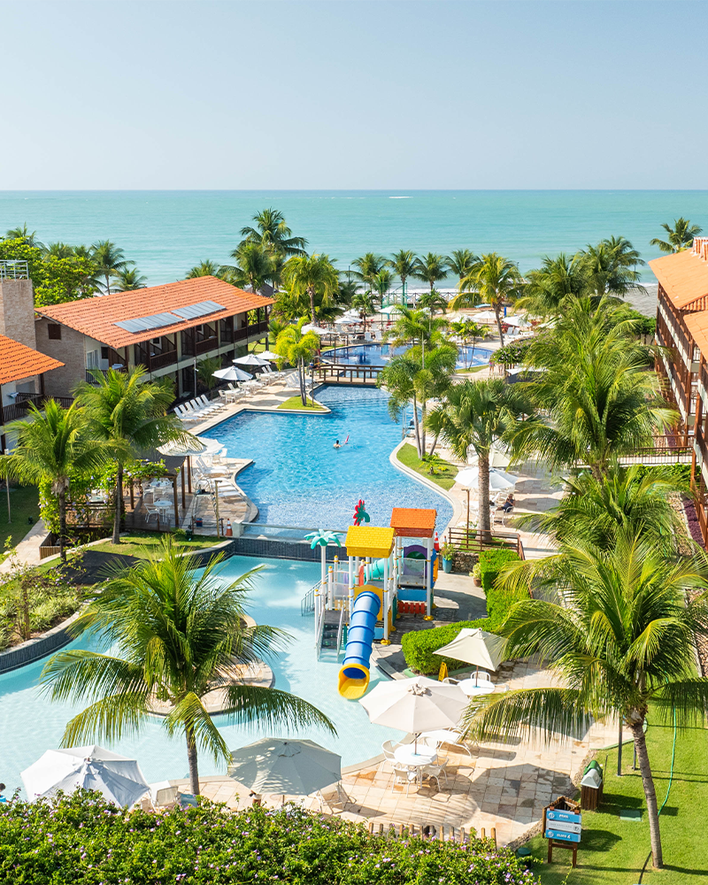 Vista aérea de um resort Salinas Maceió pé na areia com prédios de telhados laranjas. A área de lazer inclui piscinas com decks, coqueiros e um parque aquático infantil com escorregadores coloridos. O mar está visível ao fundo.