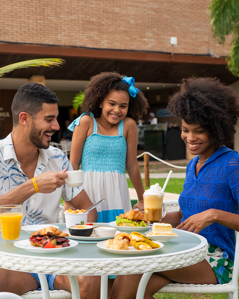Família sorrindo, sentada em mesas brancas no gramado do resort Salinas Maceió, desfrutando de um lanche com sanduíches, salgados, doces e bebidas. O pai segura uma caneca de café, a mãe uma bebida gelada e a filha observa.