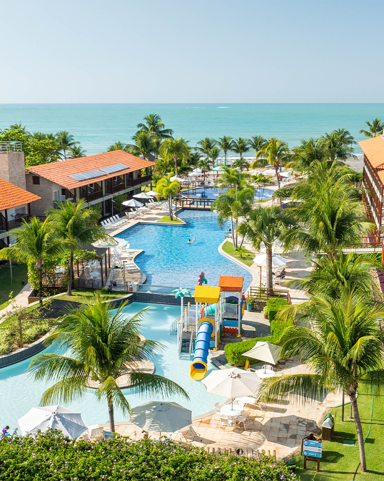 Vista aérea de um resort Salinas Maceió pé na areia com prédios de telhados laranjas. A área de lazer inclui piscinas com decks, coqueiros e um parque aquático infantil com escorregadores coloridos. O mar está visível ao fundo.