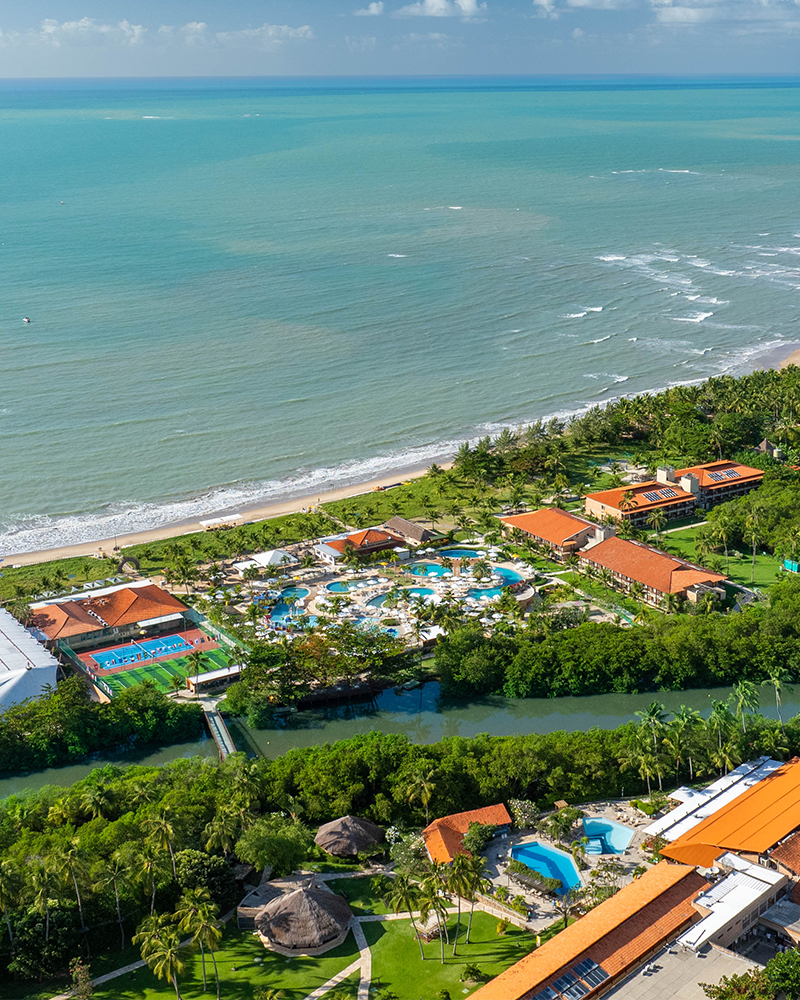 Vista de ângulo elevado do complexo do Salinas Maragogi, mostrando uma área de lazer infantil, quadras, piscinas maiores, estrutura do resort, com o rio cortando o resort com uma ponte ligando os dois lados, e o mar azul com a praia ao fundo, tudo cercado por coqueiros.