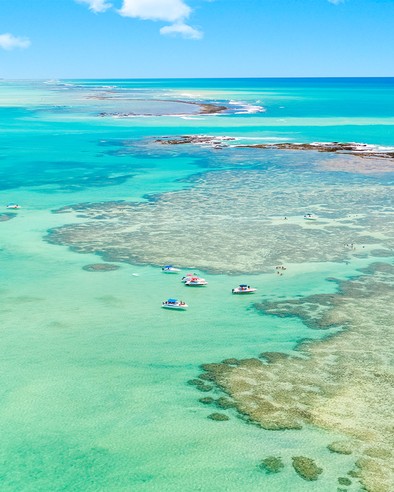 Vista aérea das piscinas naturais de Barra Grande, com águas cristalinas e formações de corais, barcos ancorados e turistas desfrutando do mar em cenário paradisíaco.
