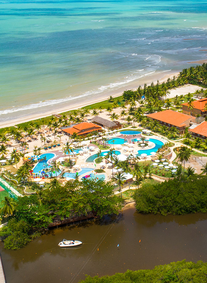 A foto mostra parte da estrutura, do ponto de vista aéreo, do Salinas Maragogi. Localizado à beira-mar de Maragogi, em Alagoas, e rodeado por mata, pode-se ver o teto de alguns de seus prédios, assim como o complexo aquático composto por várias piscinas e áreas para descanso. Um resort perfeito no nordeste brasileiro.