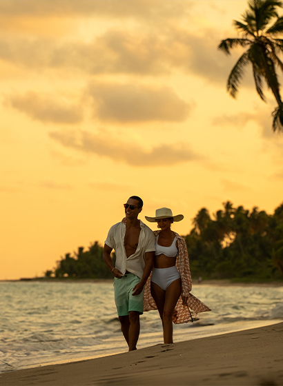 Casal caminha abraçado na beira da praia durante o pôr do sol dourado.