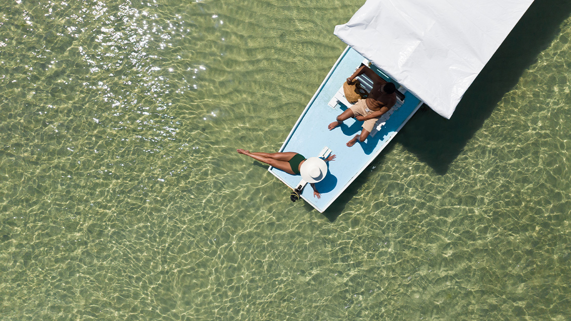 Vista aérea de um barco azul com cobertura branca navegando em águas rasas e cristalinas. Um casal relaxa a bordo enquanto o sol reflete na transparência do mar.