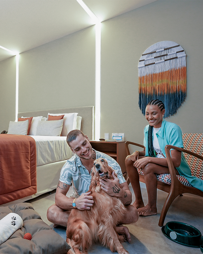 casal dog friendly Casal na Garden Exclusive com cachorro no quarto, o homem está sentado no chão alisando o dog, e a mulher sentada na poltrona olhando pra eles e sorrindo, o quarto é moderno com decoração colorida.