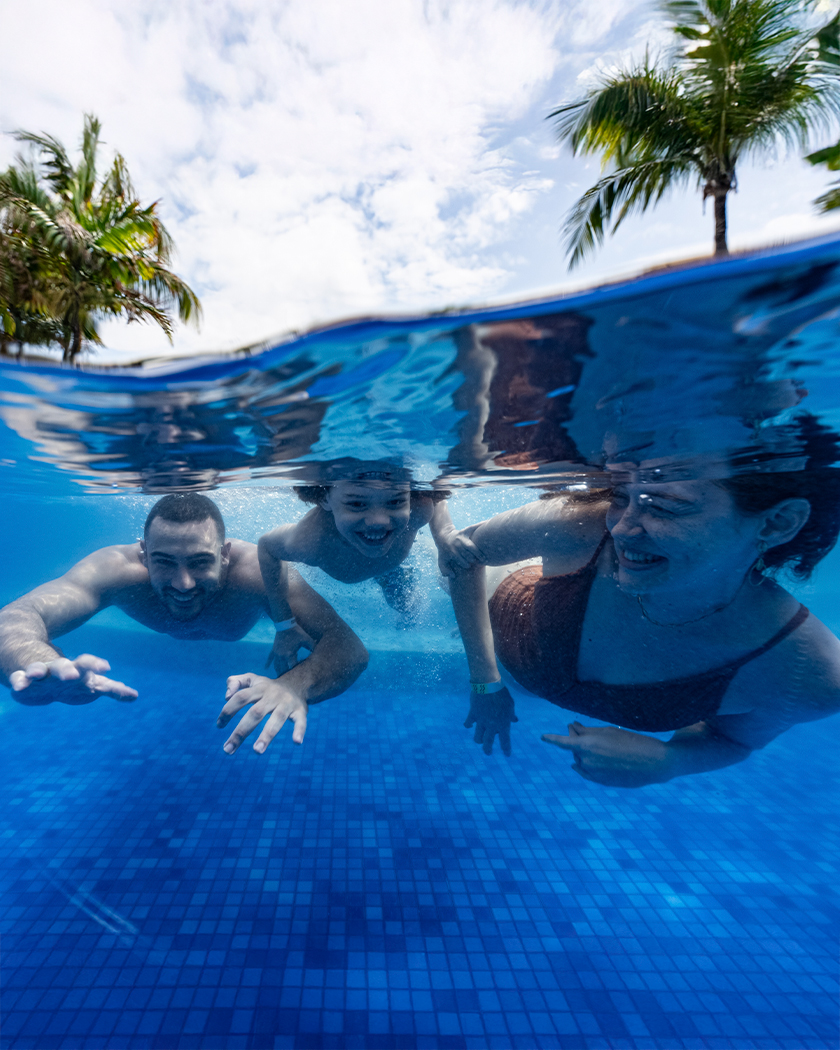 estrutura piscina Família mergulhando em uma das piscinas do Salinas Maceió, em foto subaquática, todos estão sorrindo e fora da piscina tem alguns coqueiros e o céu está nublado.