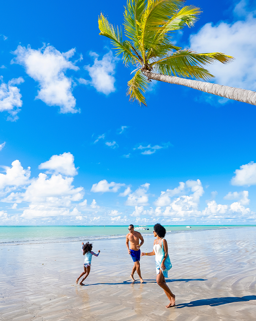 destinos paradisíacos ipioca Vista com a família correndo na areia da Praia Ipioca. A água do mar é azul-turquesa com maré baixa. O dia está ensolarado com céu azul com poucas nuvens.