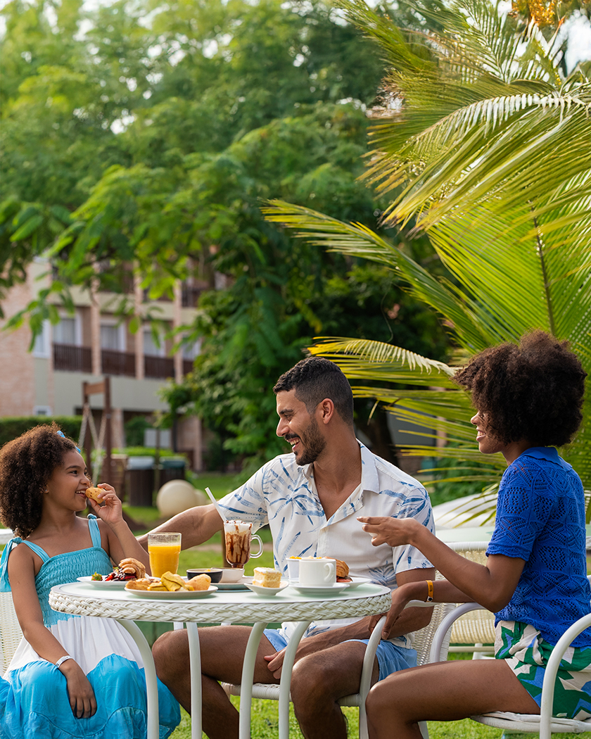petiscos all inclusive Família sorrindo e petiscando à beira da piscina em um dia ensolarado. A mesa redonda está cheia de pratos com bolos e pães diversos. O pai tem um café gourmet e a mãe está com um café simples, e sentado com um suco amarelo. Ao fundo, coqueiros e a estrutura do resort Salinas Maceió.