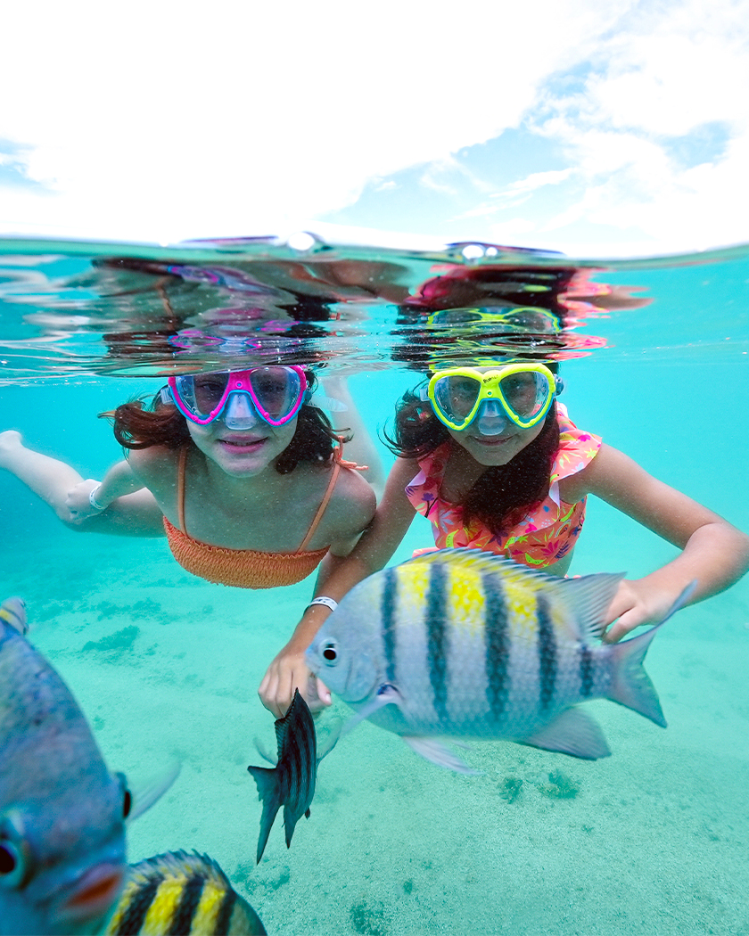 mergulho nas piscinas naturais Foto meio submersa com duas meninas usando máscara de snorkel, rodeada por peixes listrados em águas cristalinas. O fundo de areia branca é visível, e a superfície da água reflete o céu azul com nuvens.