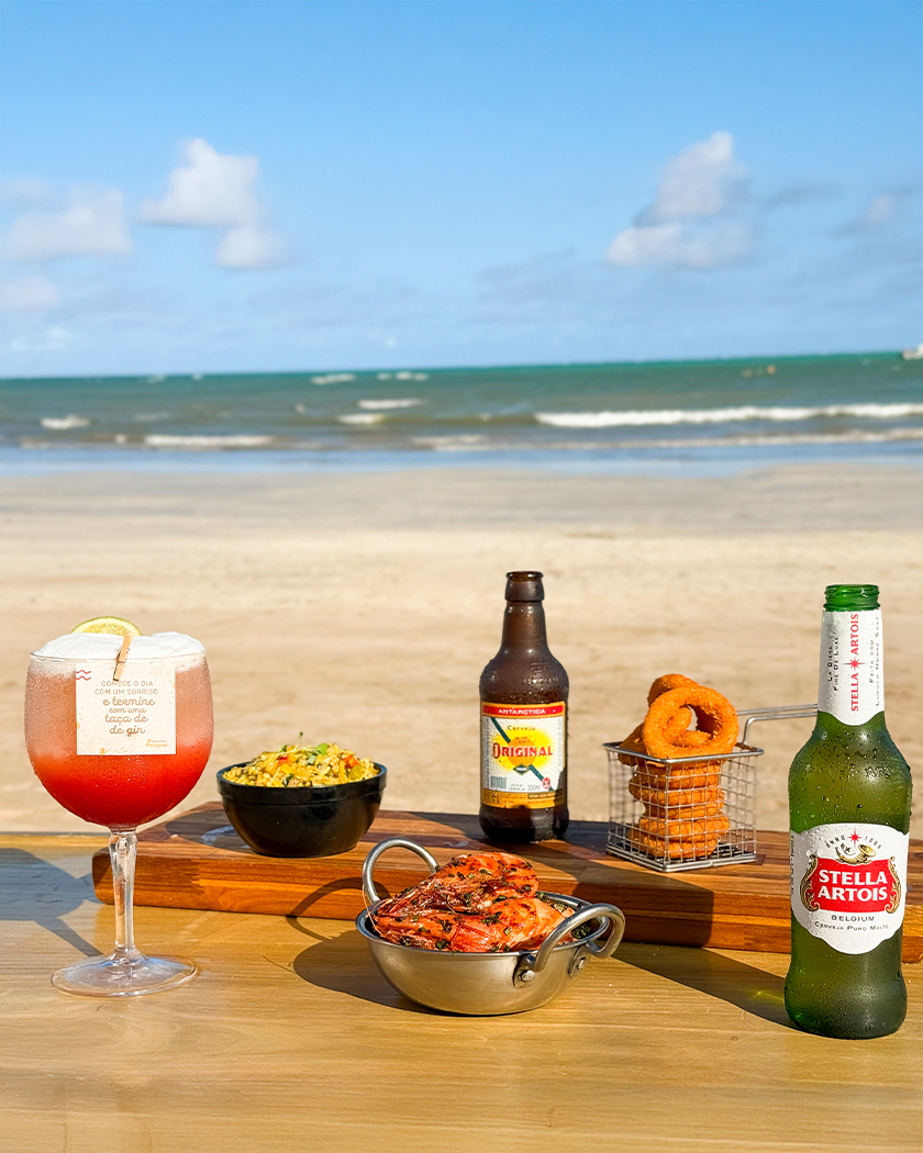 all inclusive drinks petiscos Drink vermelho, cervejas Original e Stella Artois com petiscos sendo eles camarões, onion ring, numa mesa na areia da praia, e ao fundo o mar